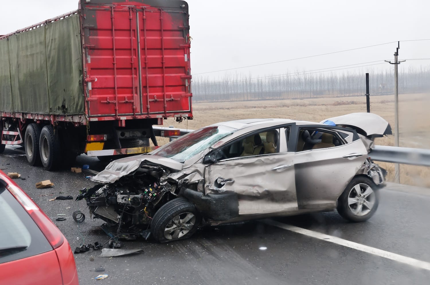 A car damaged in an accident with a semi-truck on a rainy highway.